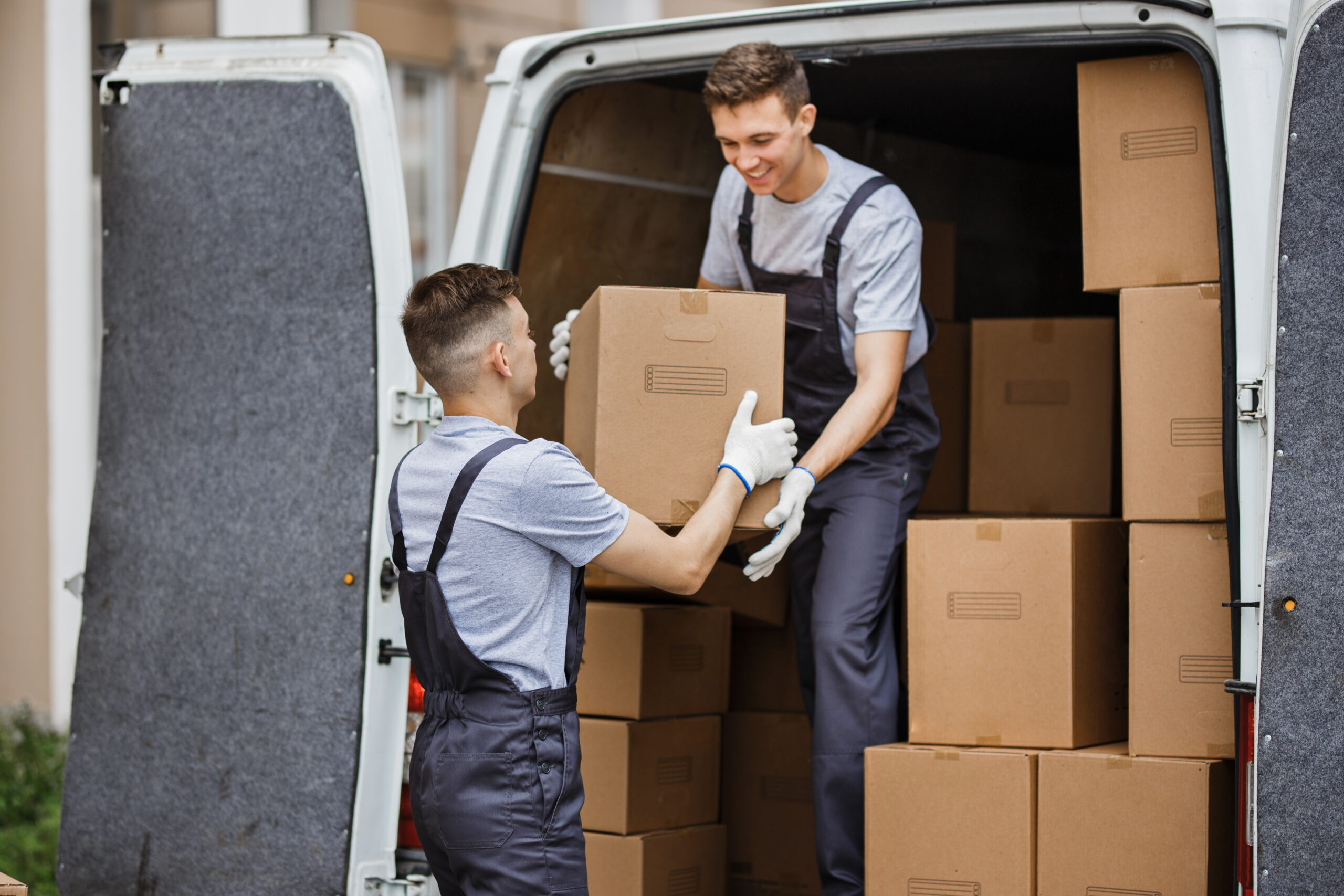 Two young handsome movers wearing uniforms are unloading the van full of boxes. House move, mover service.