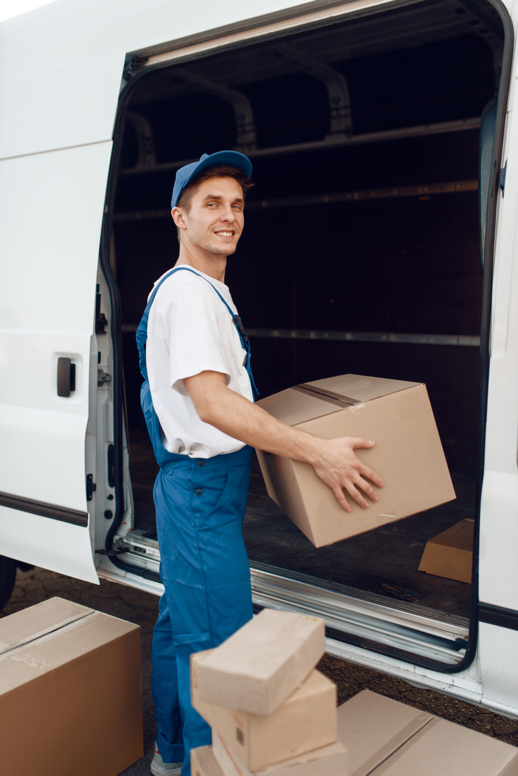 Deliveryman in uniform unloads the car with parcels, delivery service. Man standing at cardboard packages in vehicle, male deliver, courier or shipping job